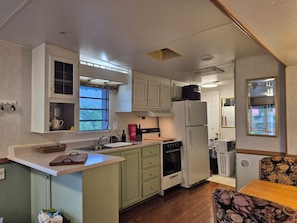A compact kitchen with a refrigerator, oven, and sink. There is a window providing natural light and a small dining area with a floral-patterned chair. - Welcome to Norma's Nest overlooking St Mary's Bay, 12kms to Digby N.S. (Digby)