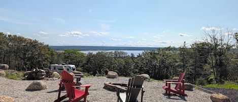 A gravel area with red Adirondack chairs and a table, a white RV, and a scenic view of a lake and mountains.