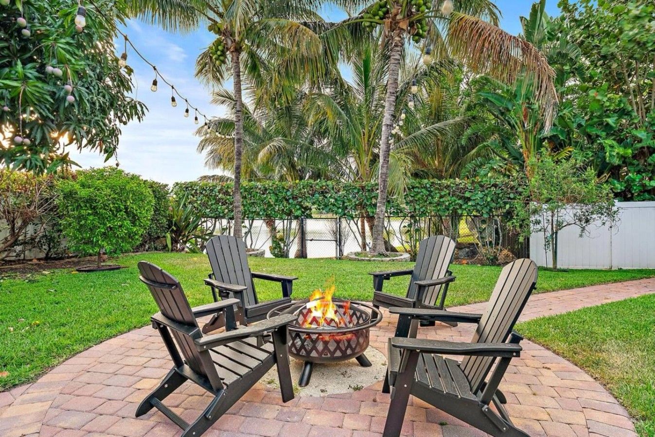A patio with four Adirondack chairs around a fire pit, surrounded by lush greenery and palm trees.