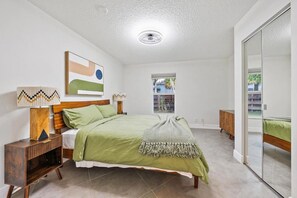 Ein Schlafzimmer mit Bett, Nachttischen, einer Lampe, einem Gemälde, einem Fenster und einer Schiebetür.