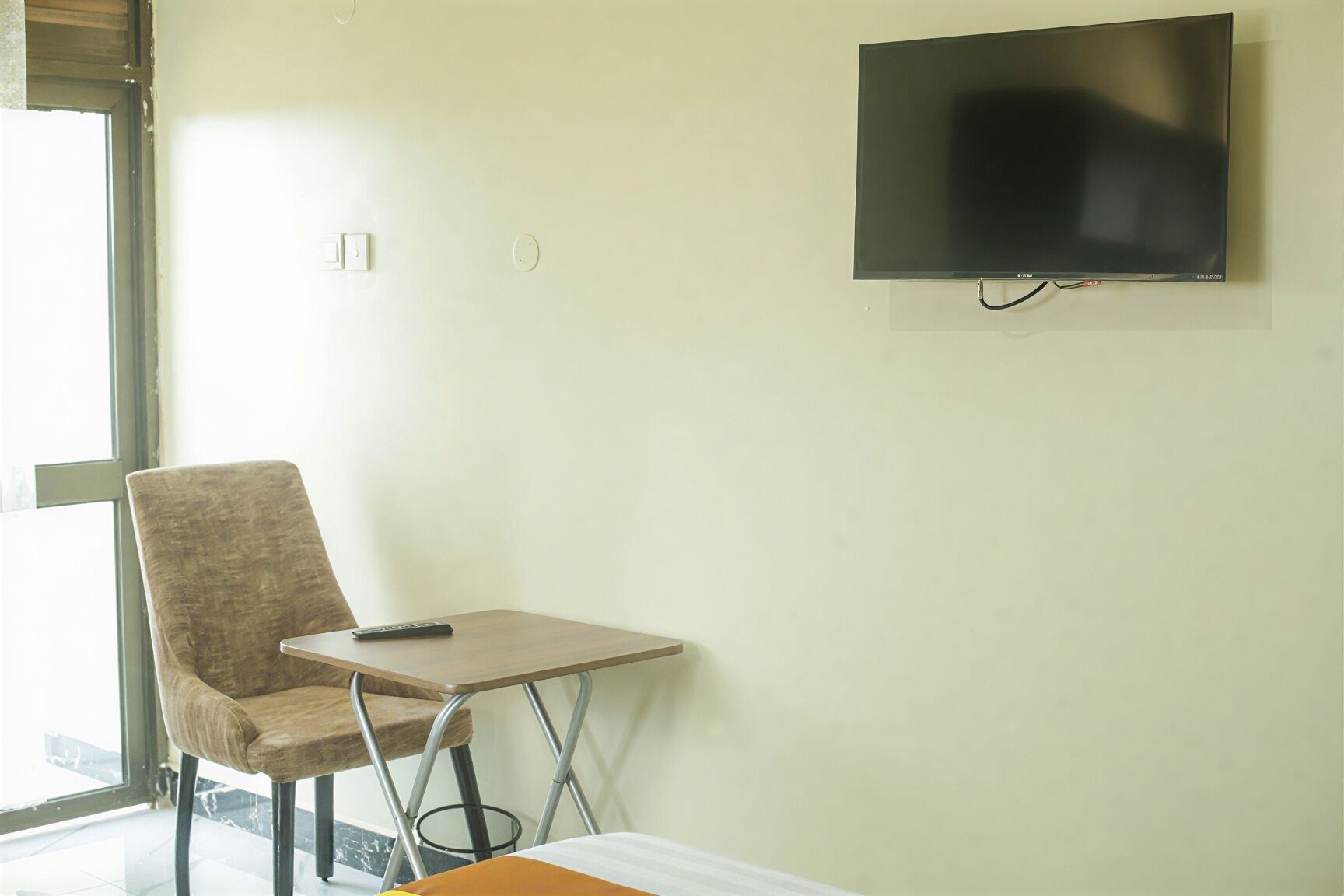 Quarto com televisão na parede, uma cadeira, uma mesa pequena e uma janela.