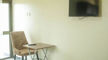 Quarto com televisão na parede, uma cadeira, uma mesa pequena e uma janela.