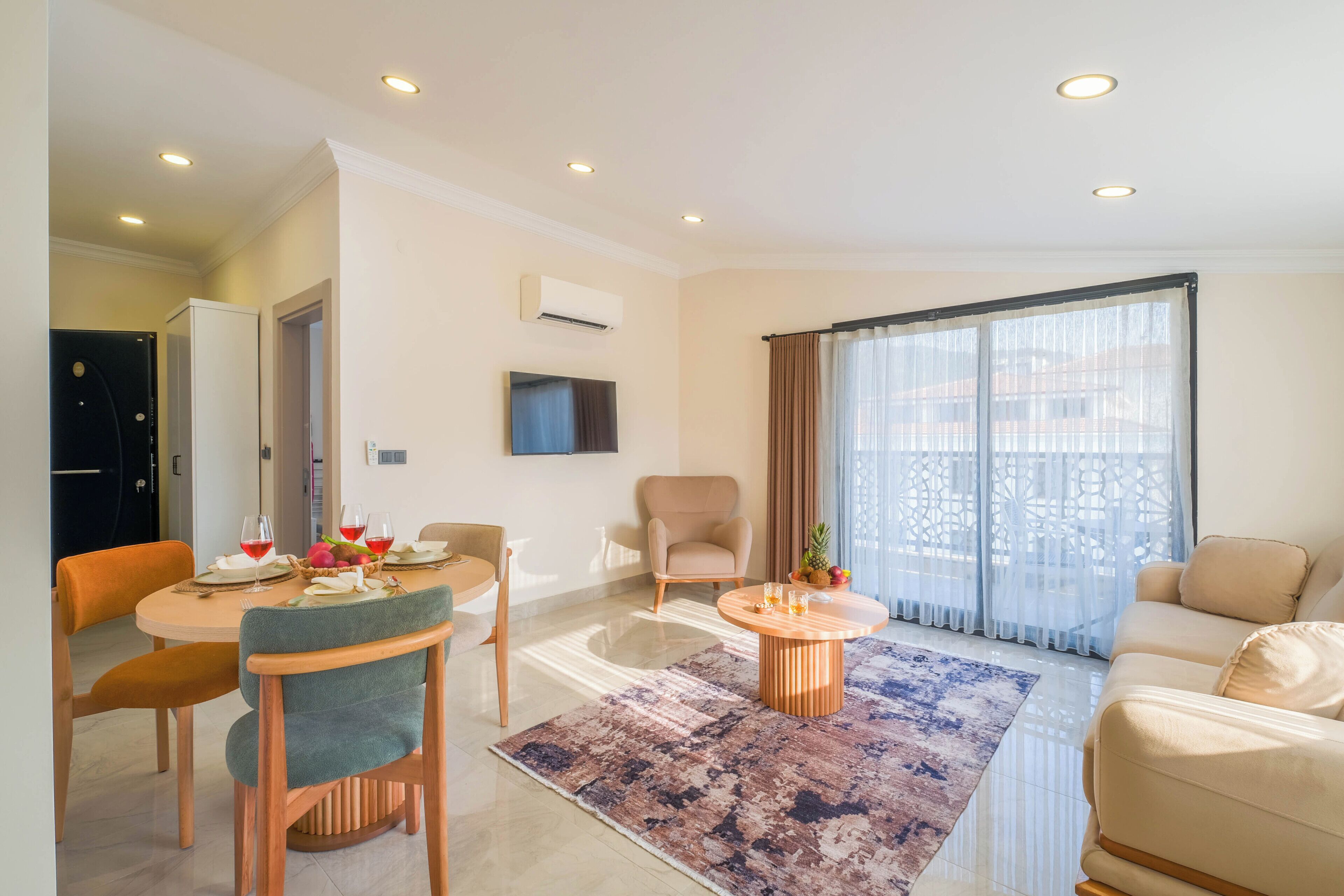 A modern living room with a dining area, a flat-screen TV mounted on the wall, and a large window with curtains.