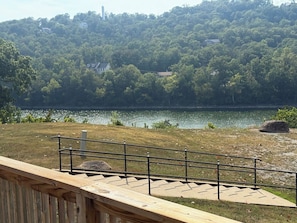A wooden deck with a metal railing overlooking a lake, surrounded by a grassy area and dense trees.