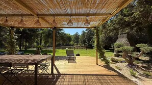 An outdoor patio with a wooden deck, a pergola with hanging lights, and a view of a garden with a stone structure. - Vrbo Property (Molac)