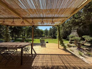 An outdoor patio with a wooden deck, a pergola with hanging lights, and a view of a garden with a stone structure. - Vrbo Property (Molac)