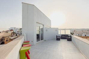 A rooftop terrace with a modern design, featuring a seating area with chairs and a table, a glass door entrance, and a view of the surrounding cityscape. - Contemporary 2BR Duplex with Sunny Terrace (Swieqi)