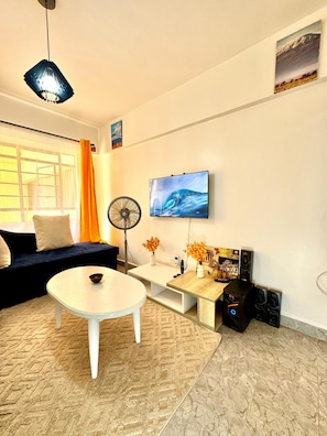 Ein Wohnzimmer mit einer Couch, einem Couchtisch, einem Fernseher, einem Ventilator und einem Computerarbeitsplatz.
