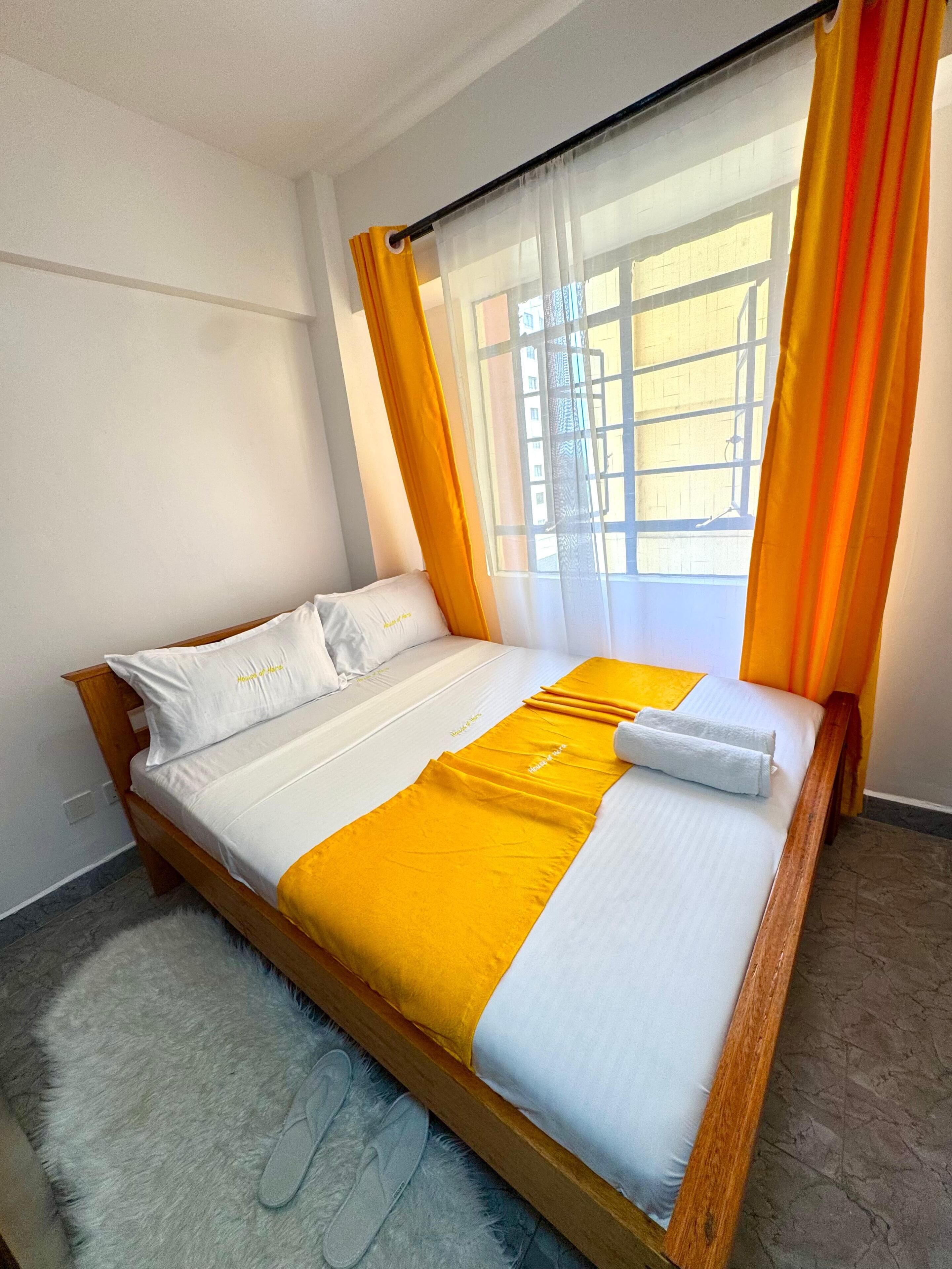 Una habitación de hotel con cama, ventana con cortinas, alfombra mullida y pantuflas.