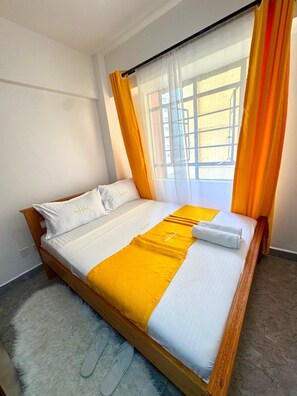 Una habitación de hotel con cama, ventana con cortinas, alfombra mullida y pantuflas.