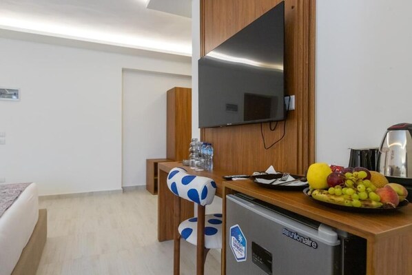 A hotel room with a bed, a desk with a refrigerator, a flat-screen TV mounted on the wall, and a dining table with a fruit bowl.