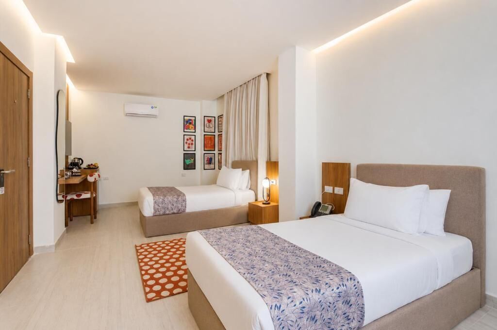 A hotel room with two beds, a desk, a chair, an air conditioning unit, and framed pictures on the wall.