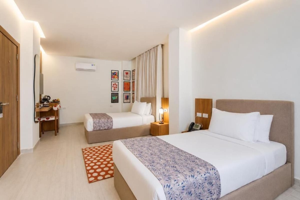 A hotel room with two beds, a desk, a chair, an air conditioning unit, and framed pictures on the wall.