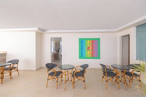 Una habitación con comedor que incluye mesas y sillas redondas, un cuadro abstracto y colorido en la pared, y vista a otra habitación a través de una puerta de vidrio.