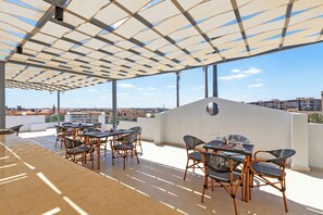 An outdoor patio area with tables and chairs, a pergola structure, and a cityscape view in the background.