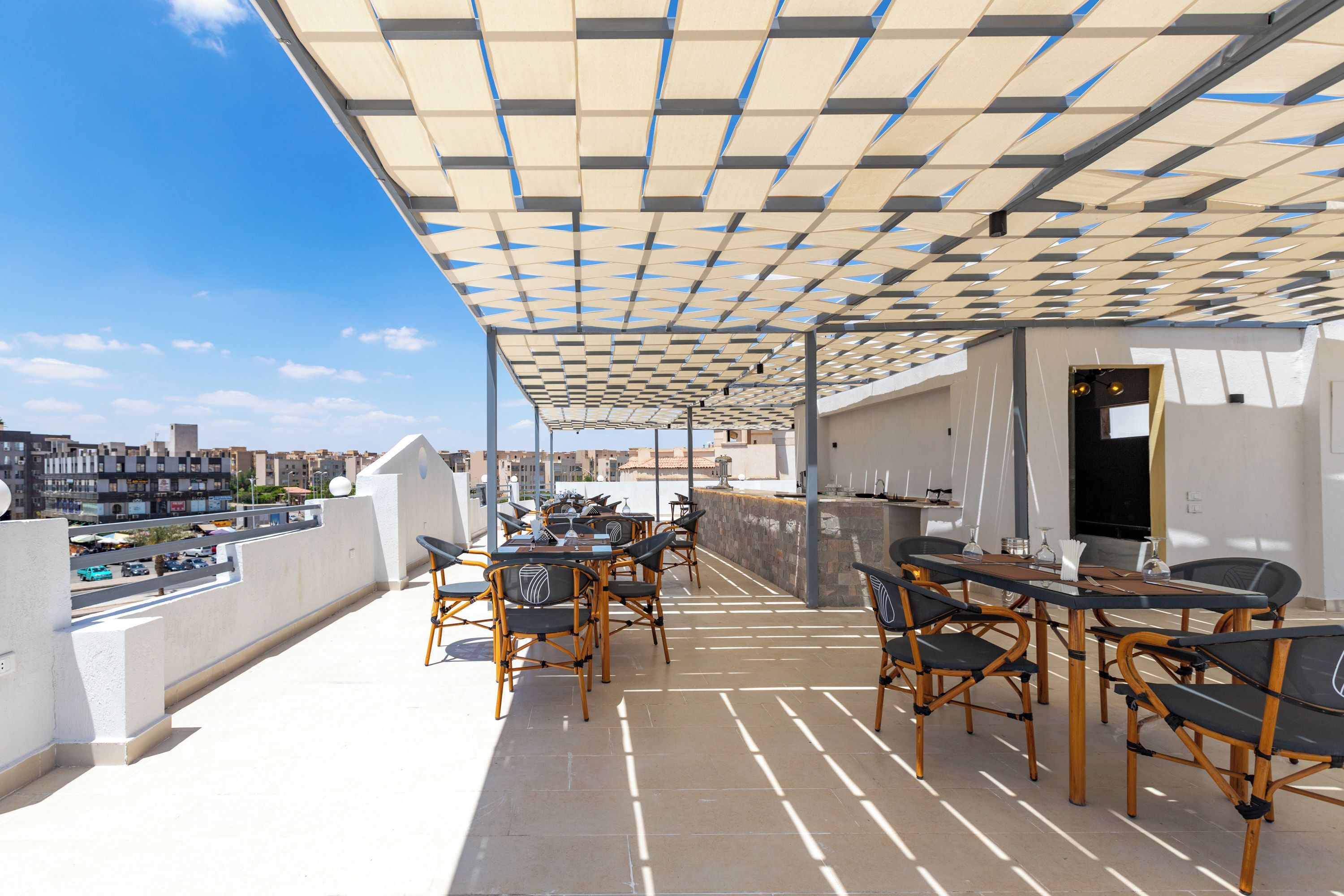 An outdoor terrace with tables and chairs, a pergola overhead, and a city view in the background.