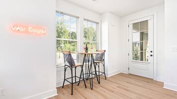 Quarto moderno com piso de madeira, paredes brancas e porta com janela. Há duas cadeiras e uma mesa com uma garrafa e copos.