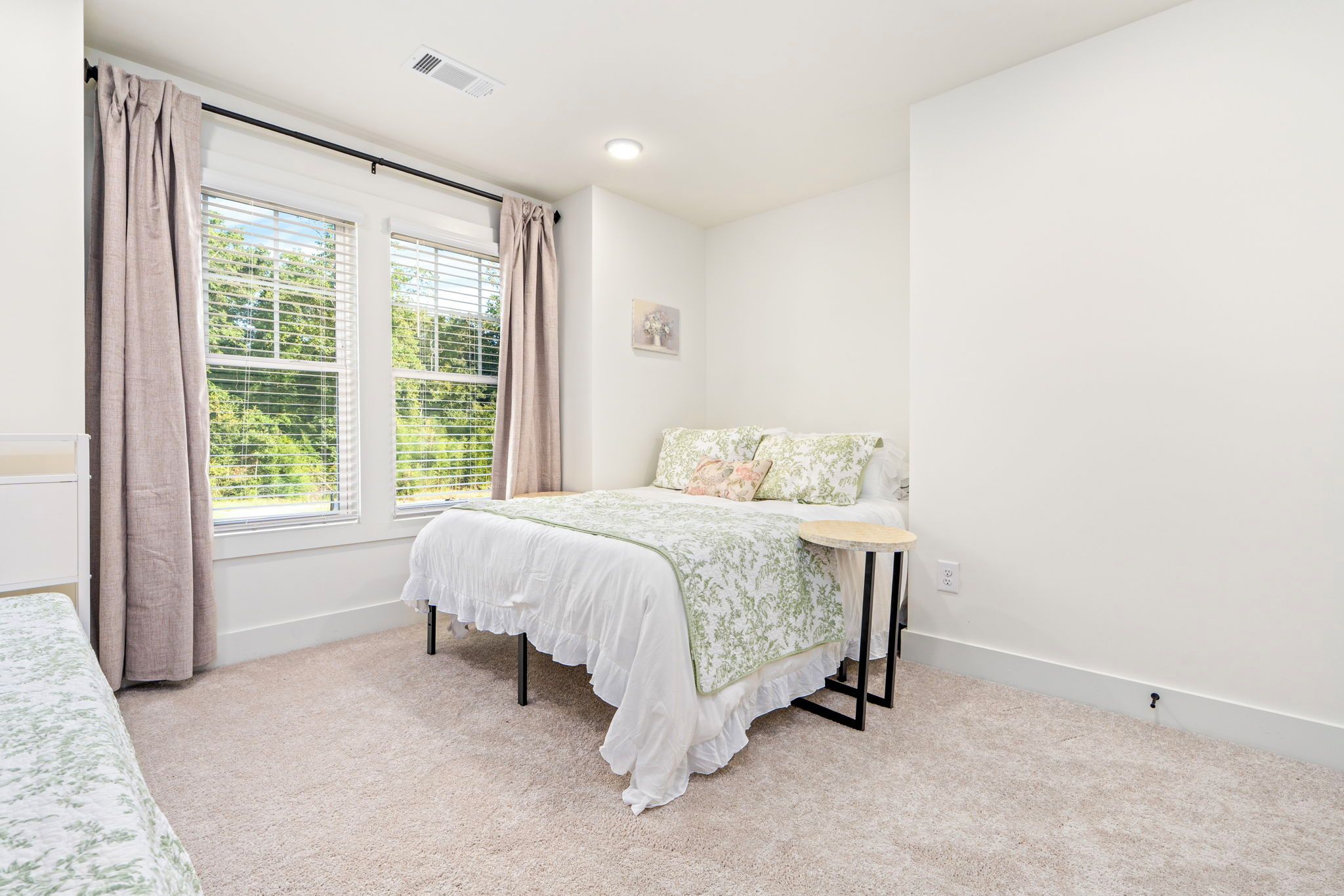 A bedroom with a bed, a bedside table, a window with curtains, and a wall-mounted light fixture.