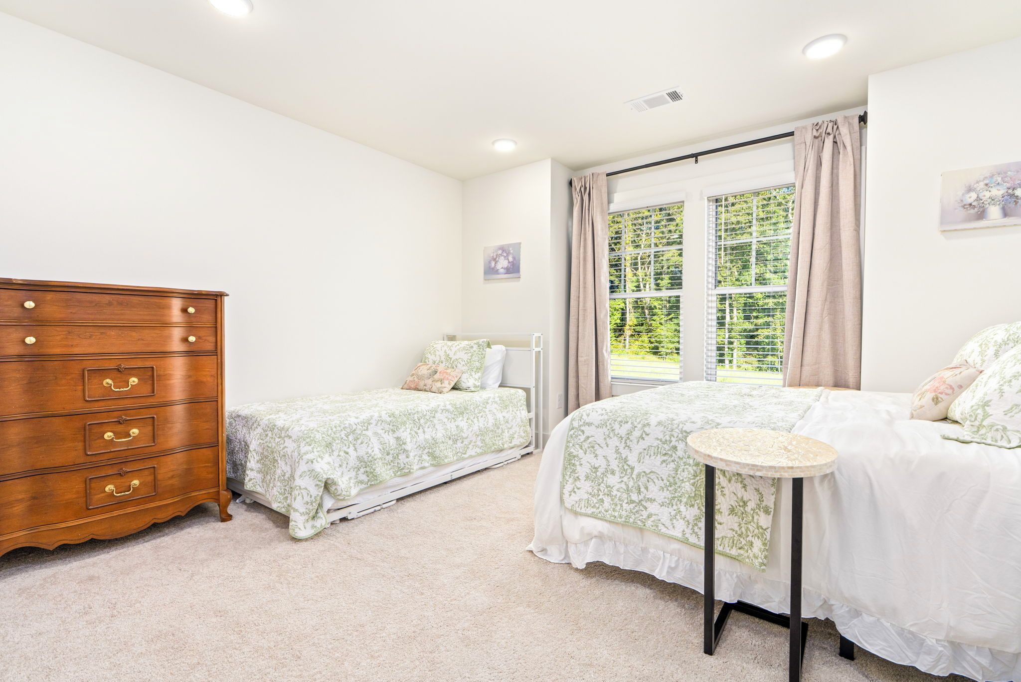 Quarto com duas camas de solteiro, cômoda de madeira, mesa redonda pequena e janela com cortinas.