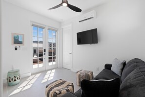 A modern living room with a ceiling fan, a flat-screen TV mounted on the wall, and a large window with a view of a building. - 🌴Calico Jack's | Steps to the Beach & Publix | Pool and Spa & Gulf Sunsets 🌅 (Fort Myers Beach)