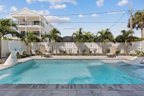 A swimming pool with a white fence, palm trees, and a two-story building in the background. - 🌴Calico Jack's | Steps to the Beach & Publix | Pool and Spa & Gulf Sunsets 🌅 (Fort Myers Beach)