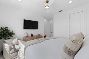 A modern bedroom with a large bed, a flat-screen TV mounted on the wall, a wooden dresser, and a potted plant. - 🌴Calico Jack's | Steps to the Beach & Publix | Pool and Spa & Gulf Sunsets 🌅 (Fort Myers Beach)