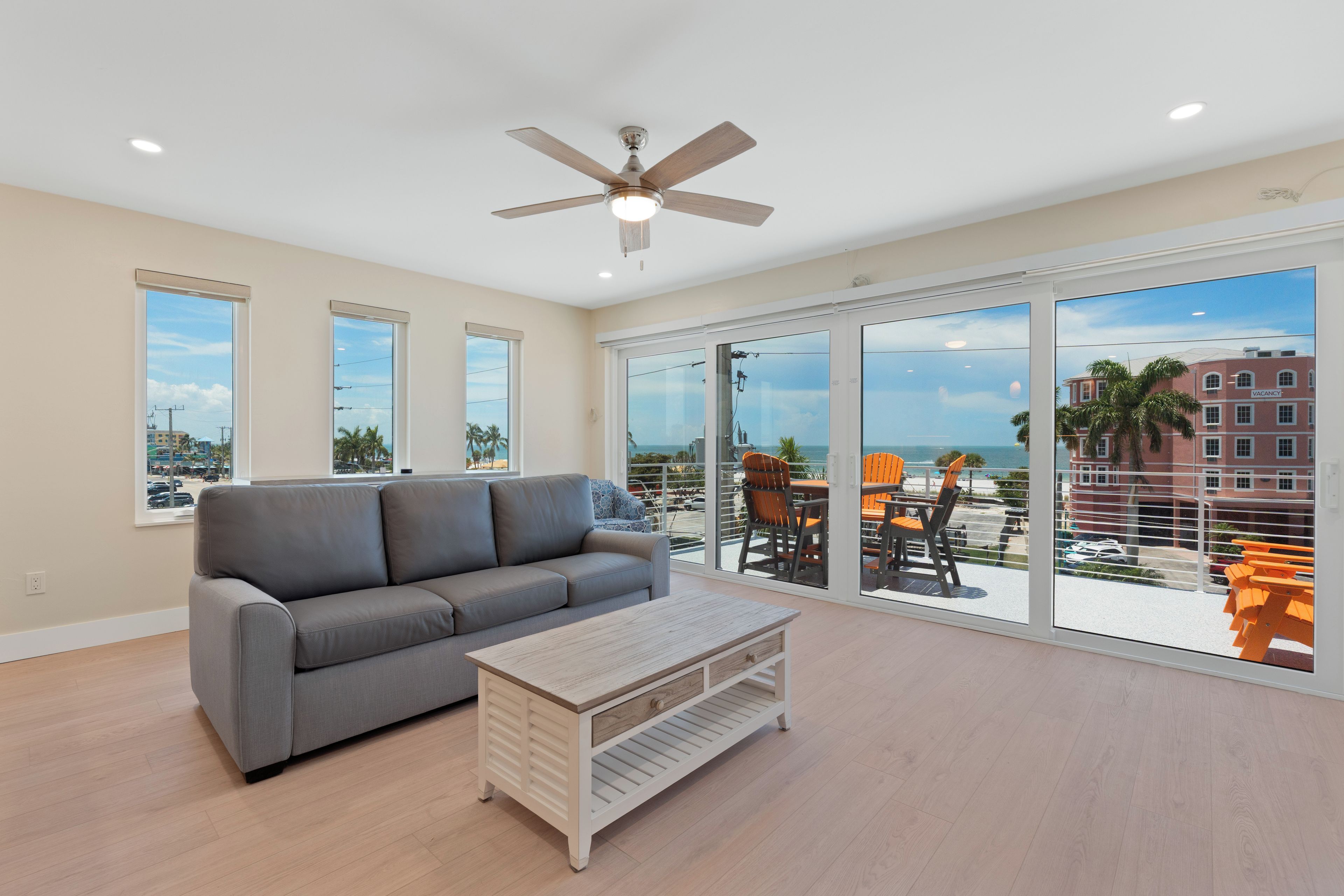 A spacious living room with a grey sofa, a wooden coffee table, and a ceiling fan. Large windows offer a view of the ocean and a building with a 'VACANCY' sign.