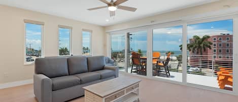 A spacious living room with a grey sofa, a wooden coffee table, and a ceiling fan. Large windows offer a view of the ocean and a building with a 'VACANCY' sign.