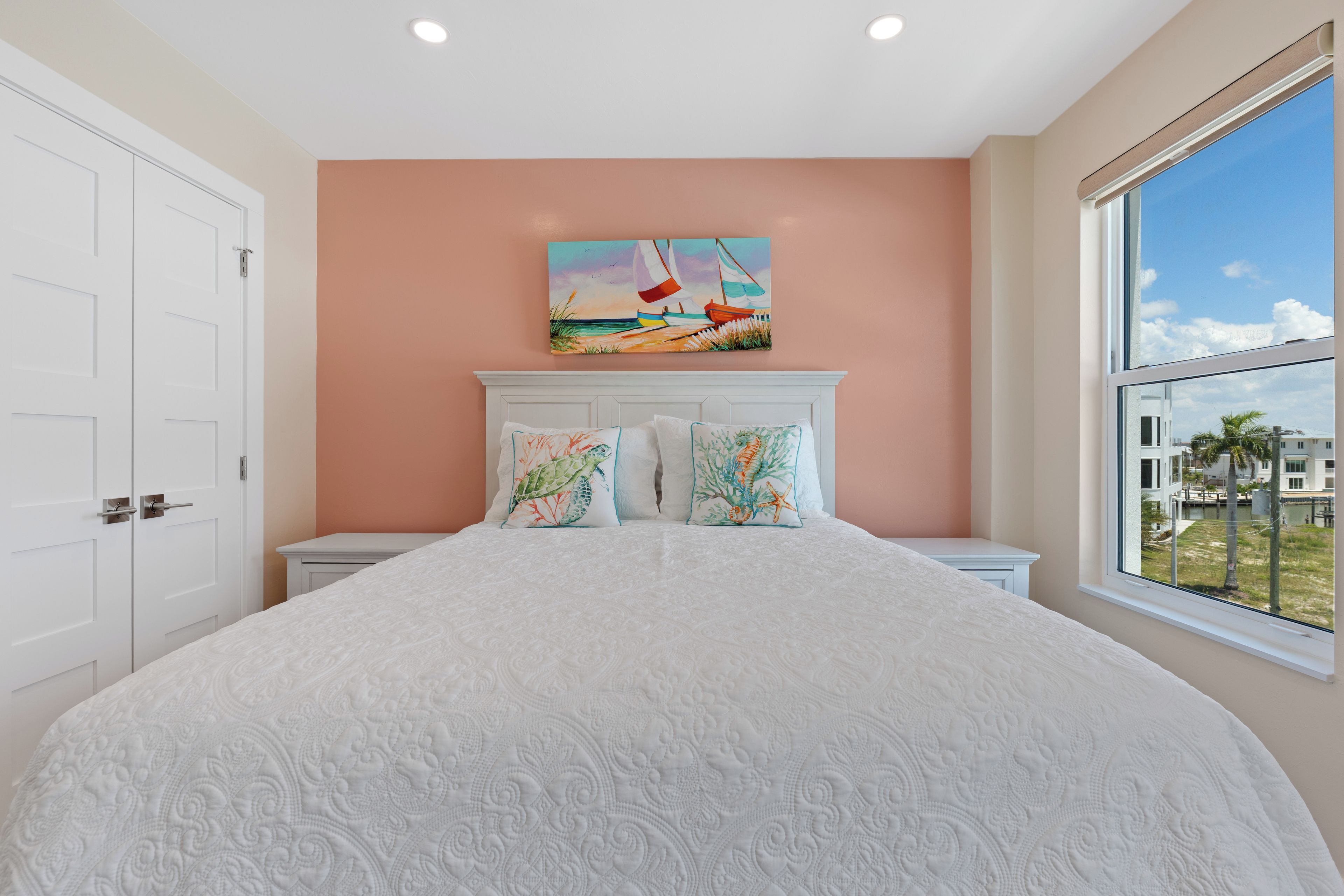 A bedroom with a large bed, two nightstands, a painting above the bed, and a window with a view of buildings and palm trees.
