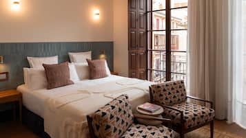 Habitación de hotel con cama, dos mesitas de noche, una silla, una pequeña mesa y ventana con vista a edificios.
