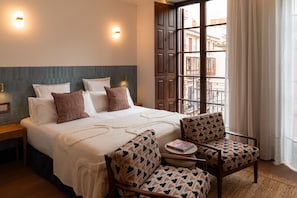 A hotel room with a bed, two bedside tables, a chair, a small table, and a window with a view of buildings.