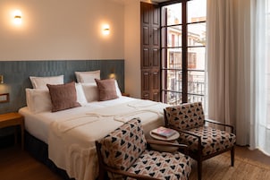 Habitación de hotel con cama, dos mesitas de noche, una silla, una pequeña mesa y ventana con vista a edificios.