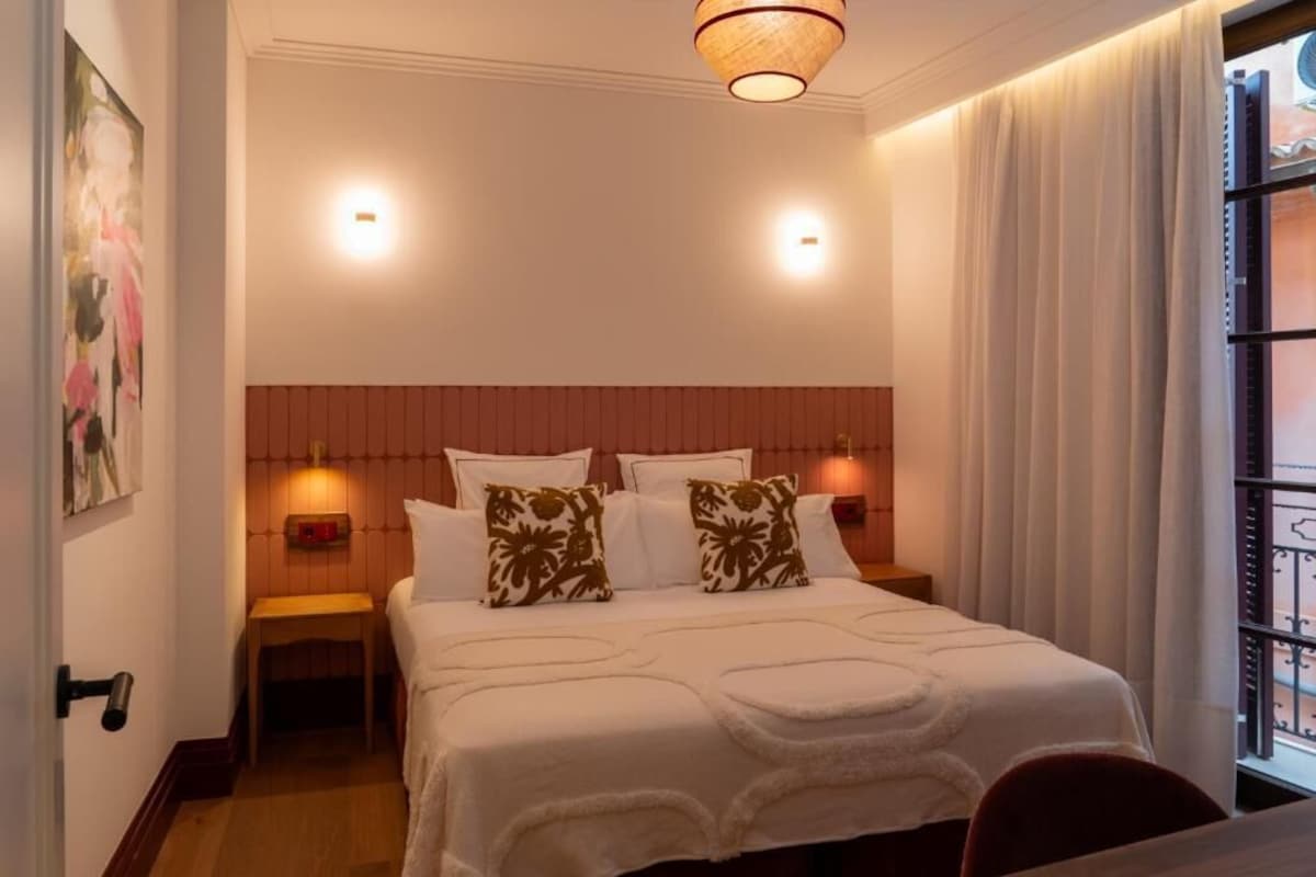 Hotellihuone, jossa on sänky, kaksi yöpöytälamppua, yöpöytä, ikkuna verhoilla ja taulu seinällä.