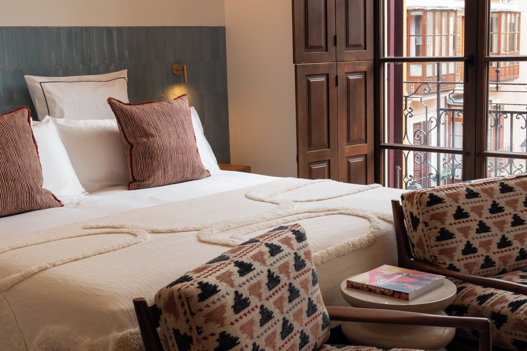 Habitación de hotel con una cama, dos almohadas, una mesita de noche con un libro y una silla con un cojín estampado.