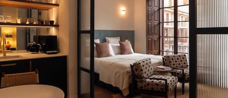 A hotel room with a bed, a sitting area with chairs, a coffee machine, and a shelf with glasses and cups.