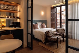 Habitación de hotel con cama, zona de estar con sillas, cafetera y estante con vasos y tazas.