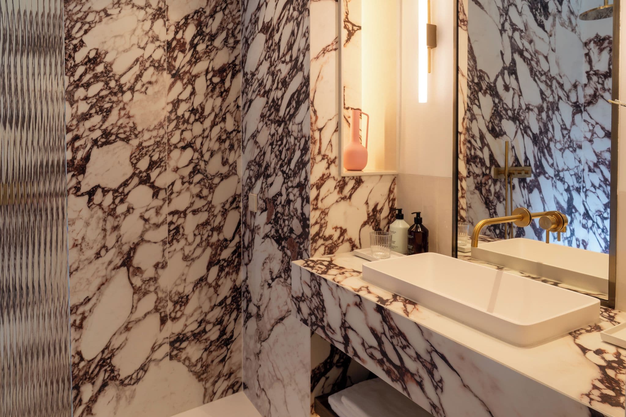 Un baño con pared de mármol, lavamanos blanco y grifo dorado.