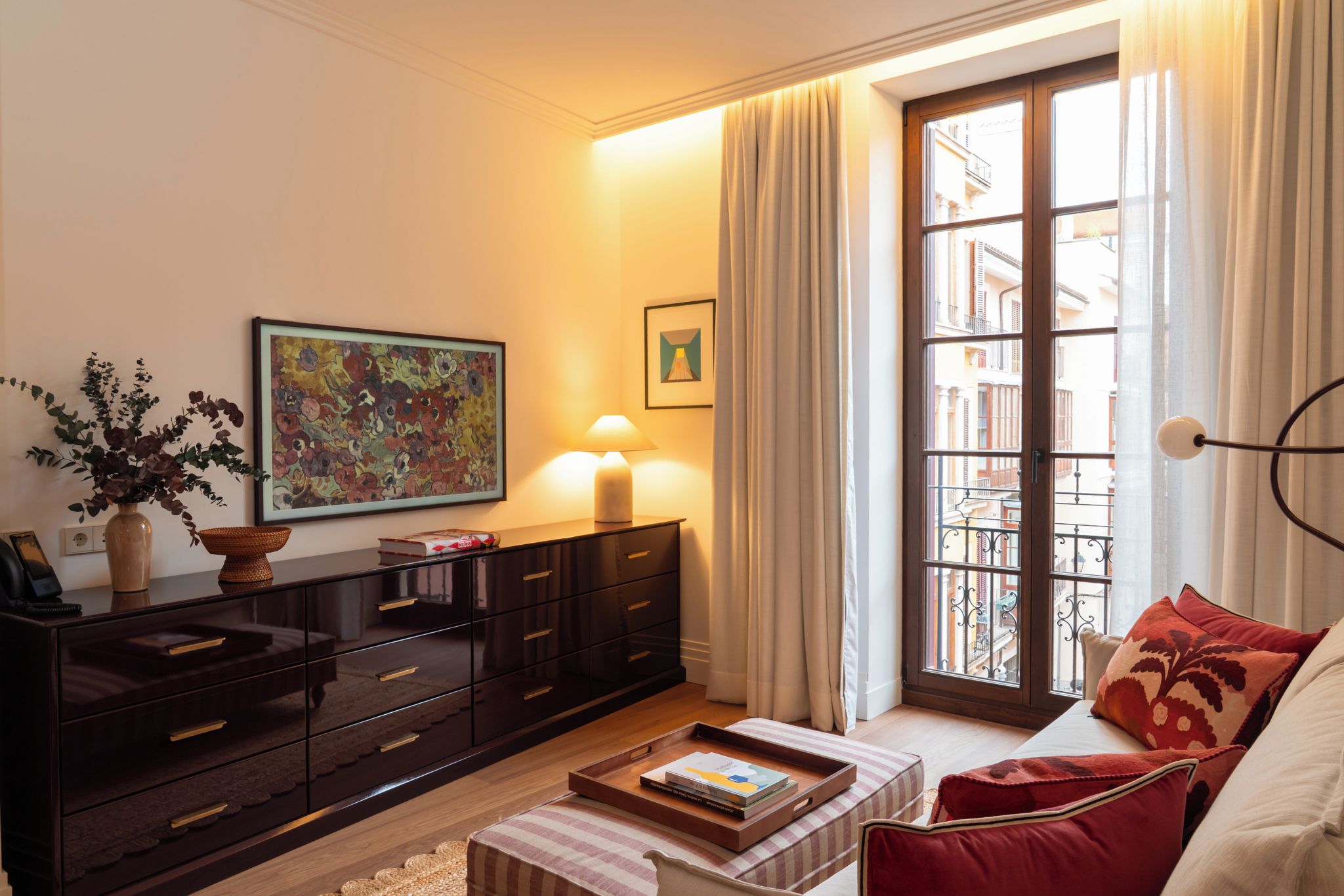 A well-lit living room with a sofa, a coffee table with books, a cabinet, a painting, and a window with curtains.