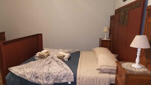 Ein Einzelbett mit blauer Bettdecke, einer beigen Decke und zwei aufgerollten Handtüchern. Dazu ein Nachttisch aus Holz mit einer weißen Lampe neben dem Bett.