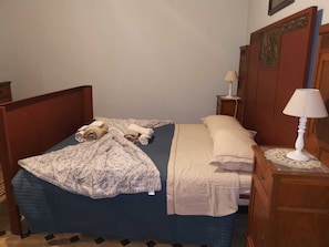 Een eenpersoonsbed met een blauwe spreideken, een beige deken en twee opgerolde handdoeken. Daarbij staat een houten nachtkastje met een witte lamp naast het bed.