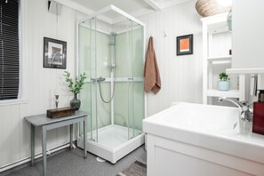 A modern bathroom with a glass-enclosed shower, a white sink, and a small table with a vase. - Cozy seaside cabin in Stamsund (Stamsund)