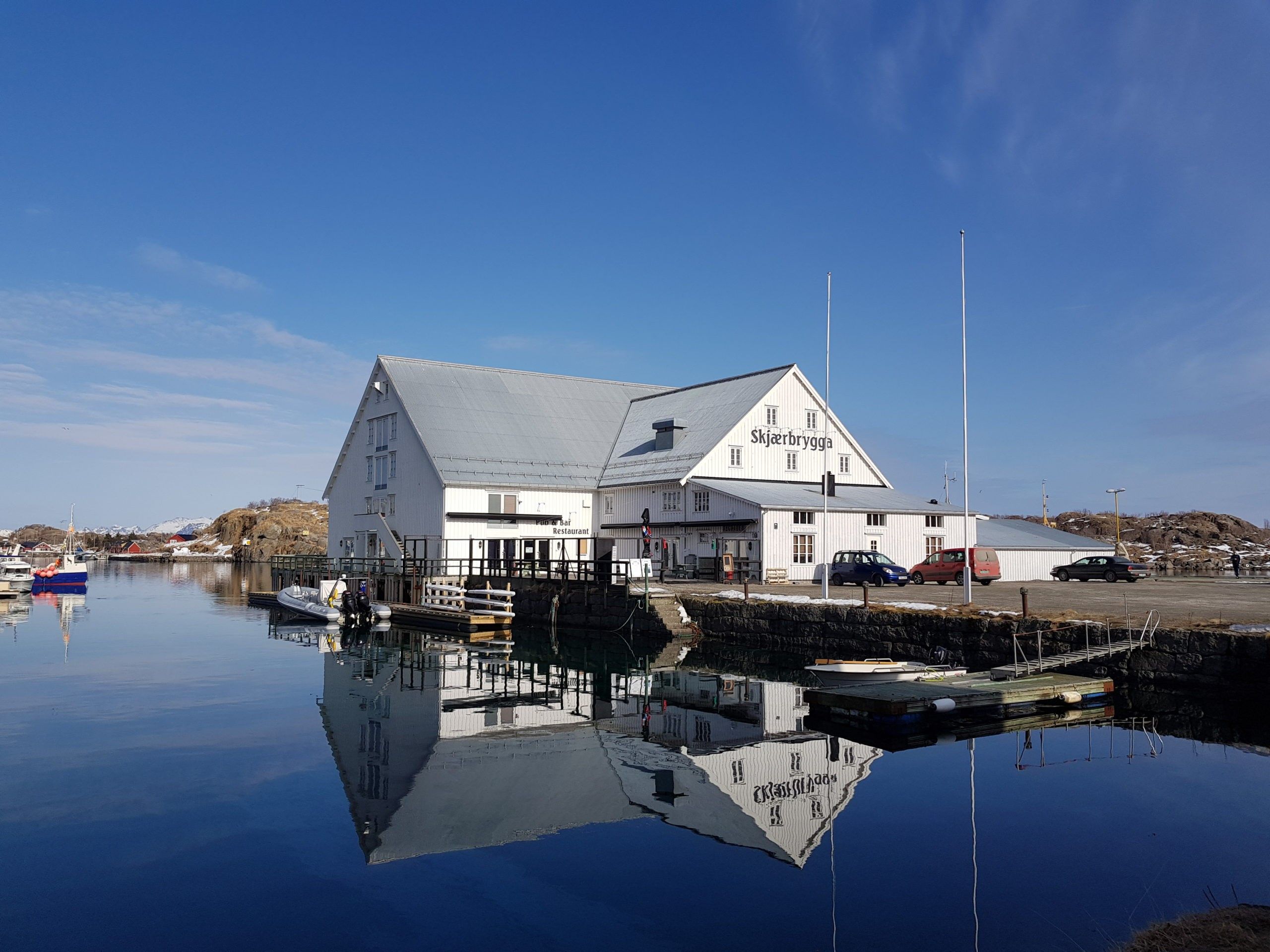A white building with a sign reading 'Skjerbrygga Restaurant' by a calm body of water reflecting the structure.