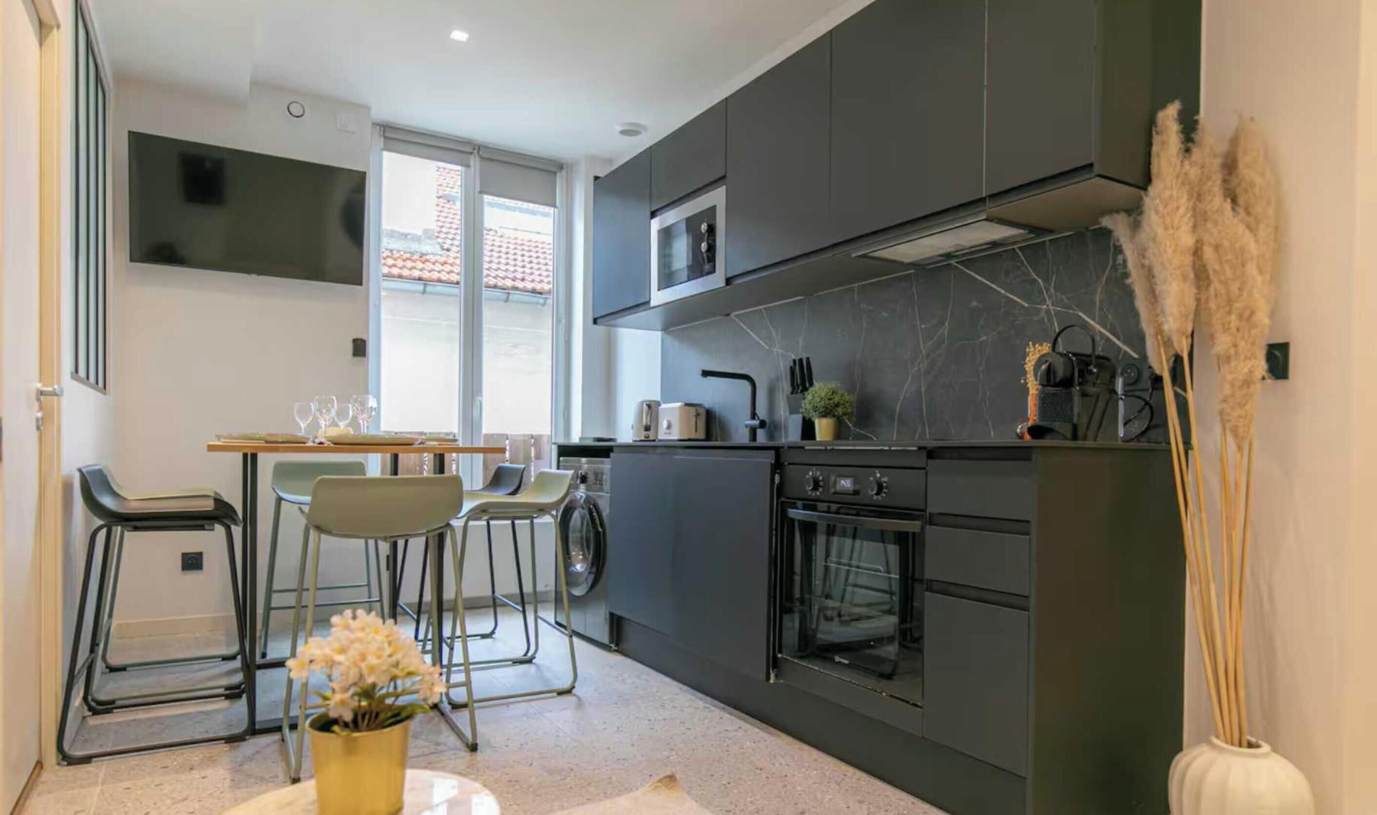 Une cuisine moderne avec des meubles de couleur sombre, un four à micro-ondes encastré et une cuisinière. On y trouve un coin repas avec des chaises et une table, ainsi qu’une fenêtre qui laisse entrer la lumière naturelle.