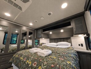 Ein modernes Schlafzimmer mit einem großen Bett, einem Einbauschrank, einem Spiegel und einem Fenster mit Blick auf Grünflächen.