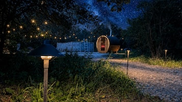 A rustic outdoor setting with a wooden structure, a metal barrel-shaped building, and a pathway leading to a lit area.