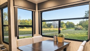 Interni di un camper moderno con tavolo da pranzo e sedute, ampie finestre che si affacciano su un paesaggio rurale.