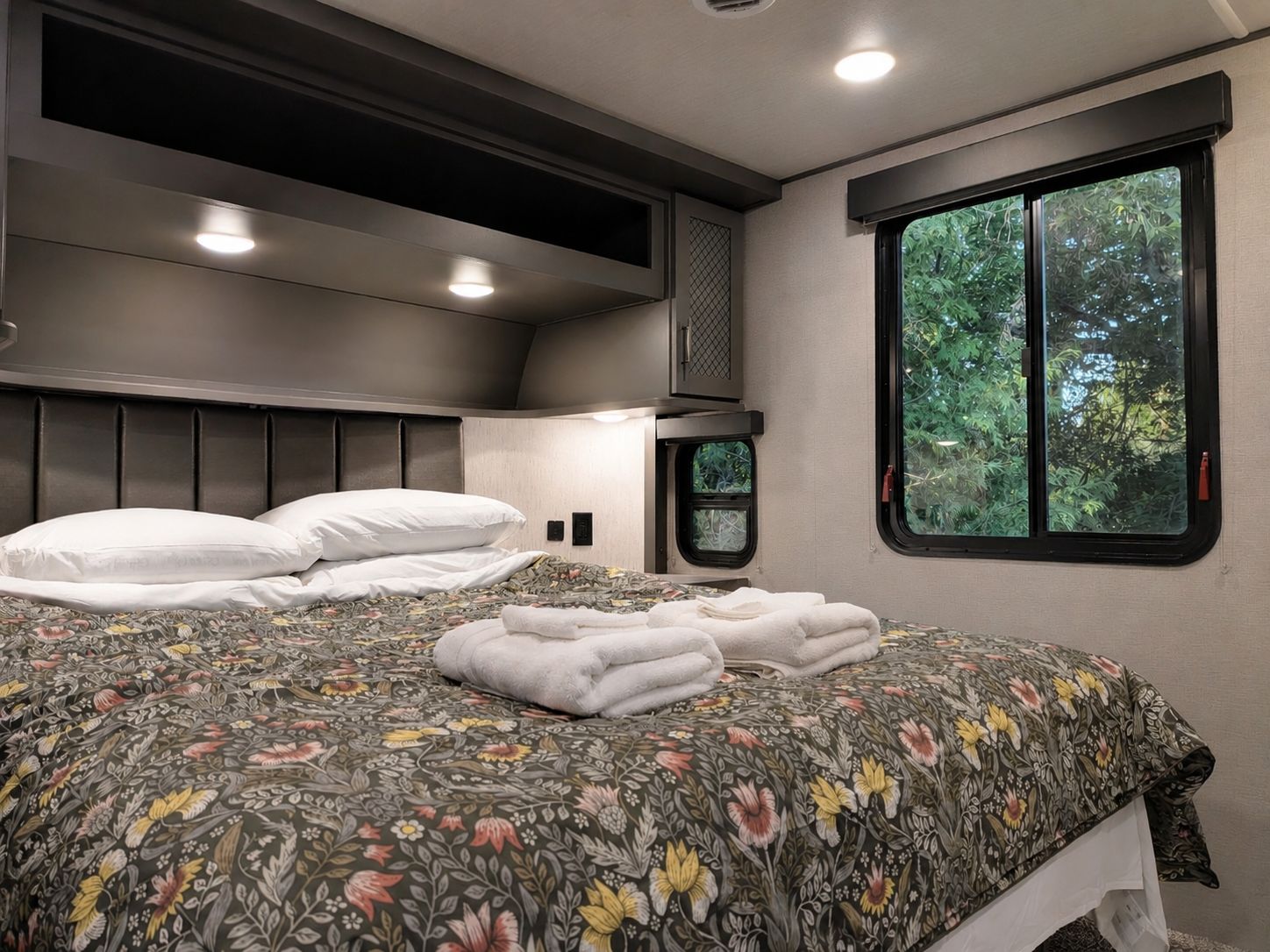 A bedroom with a bed, pillows, a window with a view of greenery, and a headboard with built-in lighting.