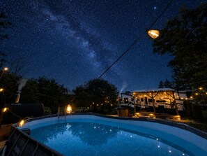 A swimming pool under a starry night sky with a house and string lights in the background.