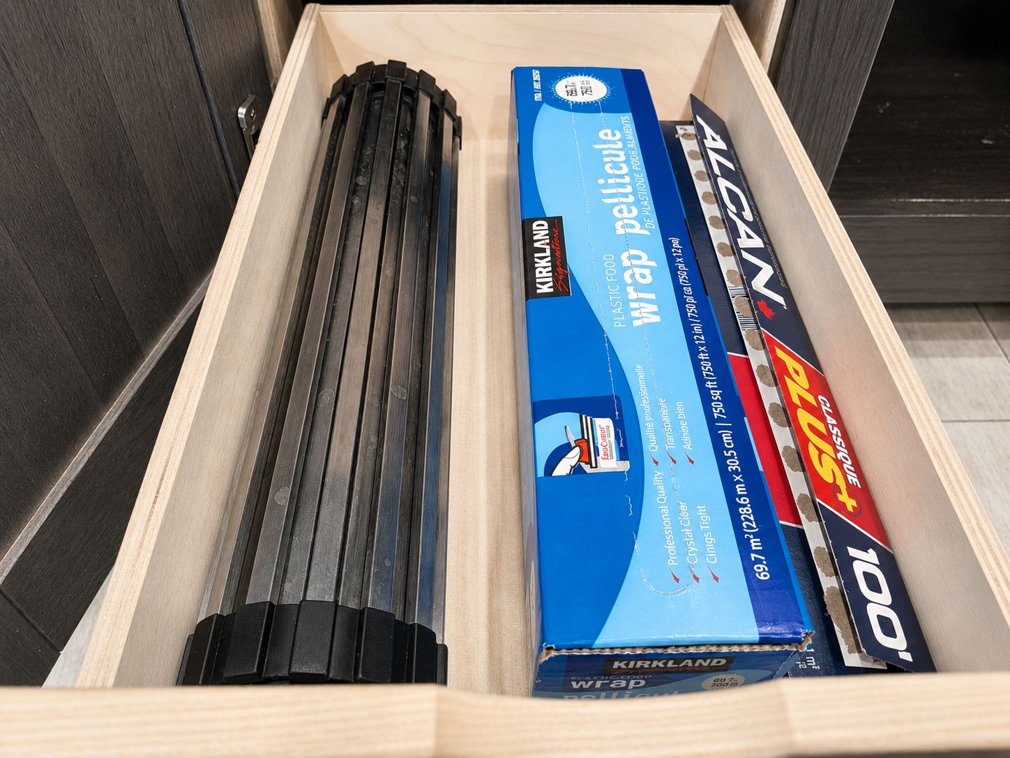 A drawer containing a black cylindrical object, a blue box labeled 'KIRKLAND Wrap,' and a red and blue box labeled 'PLUS.'