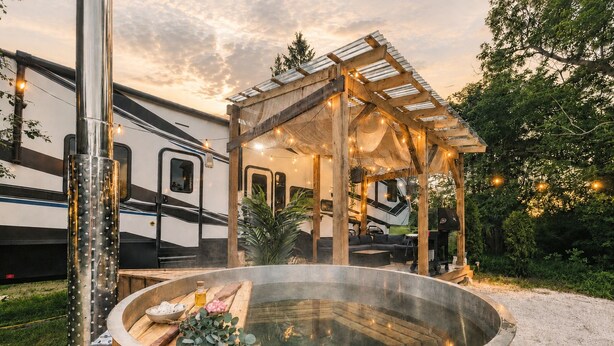 Una vasca idromassaggio con rivestimento in legno, una zona salotto coperta e un camper sullo sfondo.